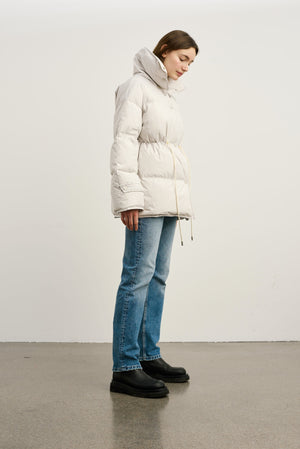 Lys, quilted jakke fra MEOTINE med justerbar talje, kombineret med blå jeans og sorte støvler, set fra siden.