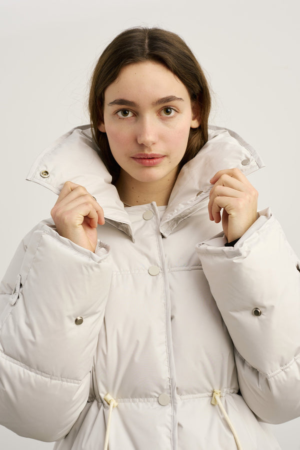 Lys, pufferjakke fra MEOTINE med høj krave, præsenteret af en ung kvinde, der holder kraven op.