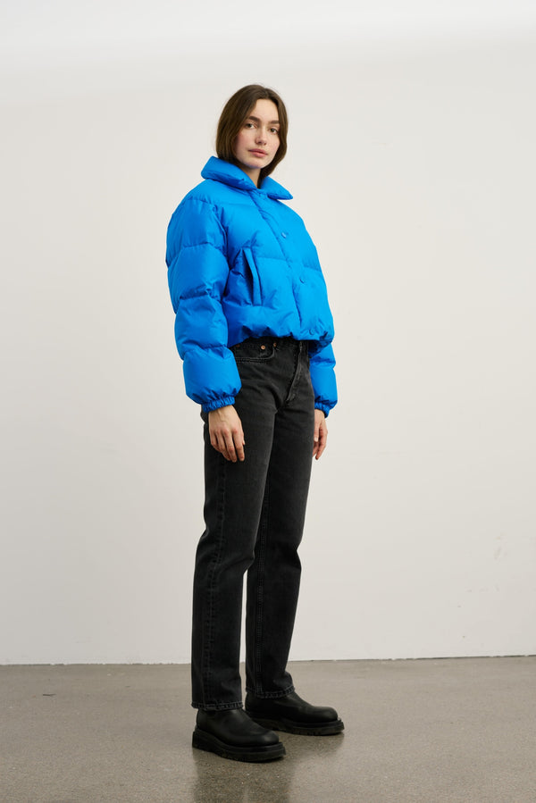 Blå, kort pufferjakke fra MEOTINE, stylet med sorte jeans og støvler, præsenteret af en model i et lyst rum.