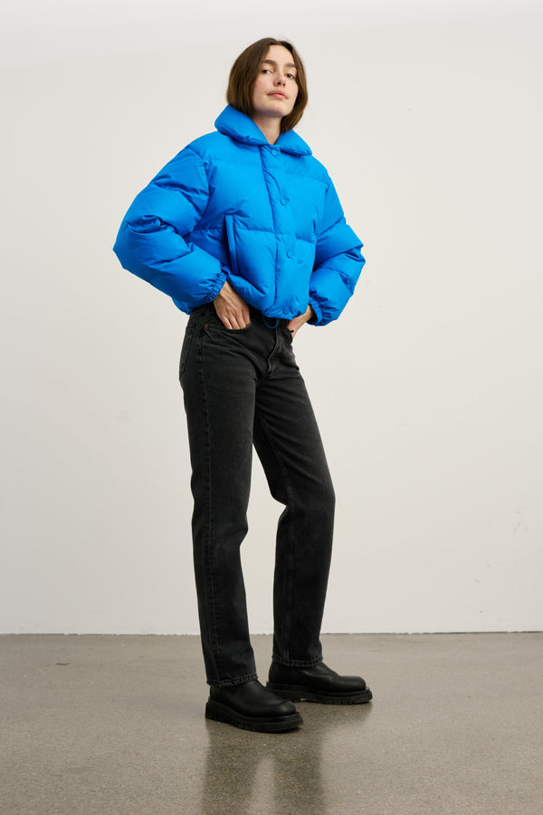 Blå, kort pufferjakke med hætte og elastiske manchetter, parret med sorte jeans og støvler. MEOTINE-stil til hverdag.