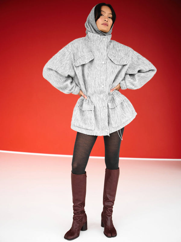 Grå jakke med hætte og store lommer, båret af model med sorte tights og brune støvler. MEOTINE tilføjer stil og komfort.