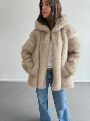 Lys beige faux fur jakke fra MEOTINE med hætte, båret af en kvinde iført en hvid top og jeans, står i et lyst rum.