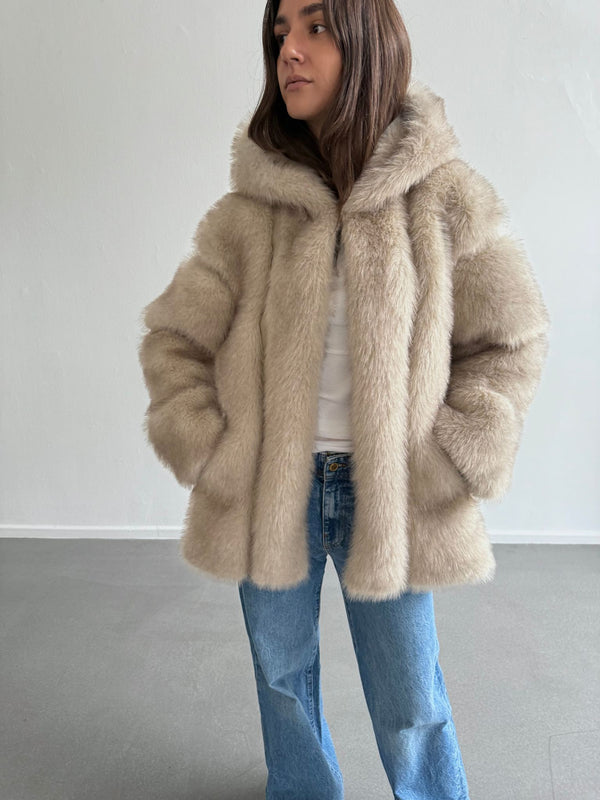 Lys beige faux fur jakke fra MEOTINE med hætte, båret af en kvinde iført en hvid top og jeans, står i et lyst rum.