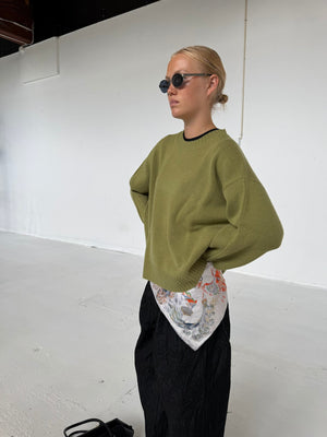 Ung kvinde iført en grøn sweater og sorte bukser, med et farverigt tørklæde fra MEOTINE knyttet om livet.