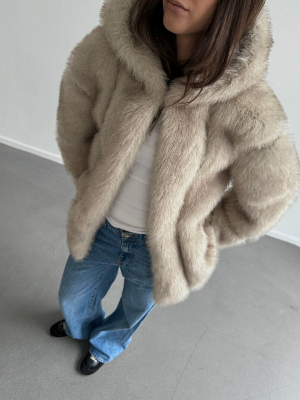 Lys beige faux fur jakke fra MEOTINE med hætte, båret af en kvinde iført en hvid top og jeans, står i et lyst rum.