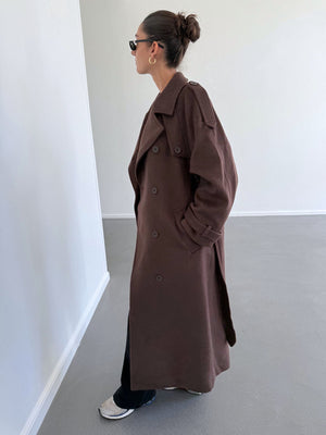 Brun trenchcoat fra MEOTINE, båret af en model med solbriller, der står i en lys, minimalistisk indendørs setting.