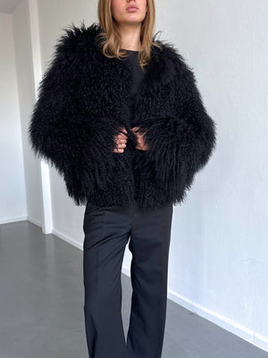 Sort, fluffy jakke fra MEOTINE med brede ærmer, bæres over en sort top og bukser, i en lys, moderne indendørs setting.