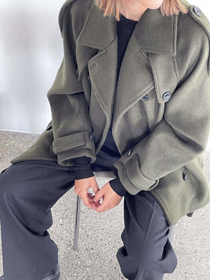 Stilfuld oversize frakke i olivengrøn fra MEOTINE, båret af en person, der sidder på en stol med sorte bukser.