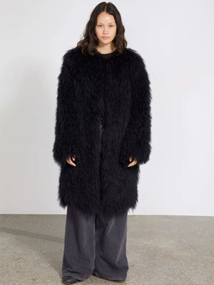 Lang, sort pelsjakke fra MEOTINE med en fluffy tekstur, båret af en model i en minimalistisk indstilling.