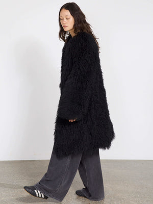 Sort, fluffy frakke fra MEOTINE, der falder løst over en model iført grå bukser og sorte sko, med en minimalistisk baggrund.