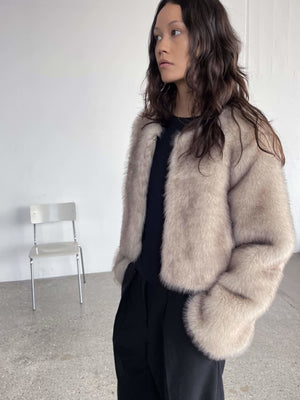 Lys beige faux fur jakke fra MEOTINE, med kort snit og bløde ærmer, båret af en model i en minimalistisk indretning.