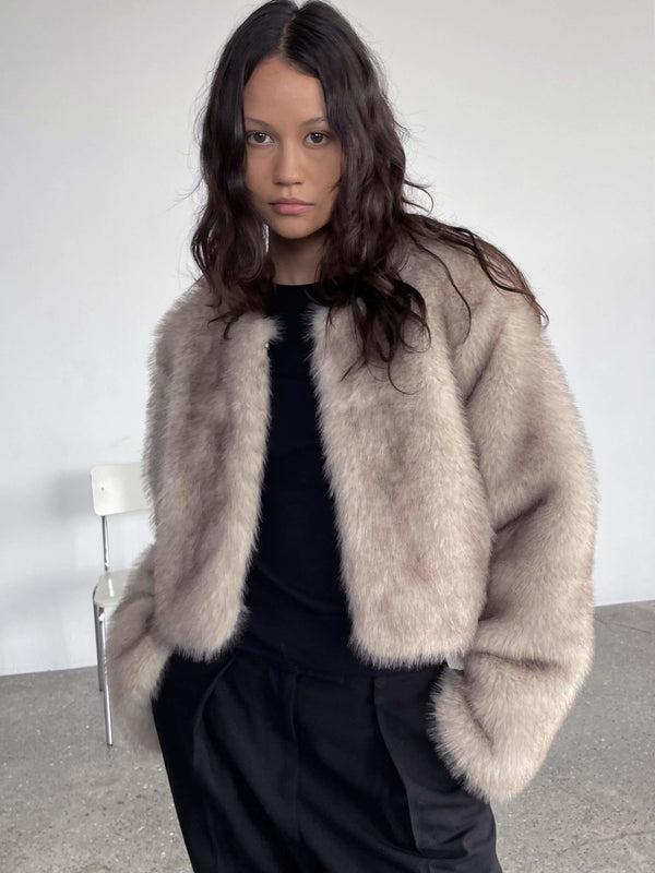 Lys, fluffy jakke fra MEOTINE, bæres af model med bølget hår, iført sort top og sorte bukser, i minimalistisk rum.