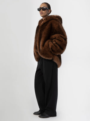 Brun pelsjakke fra MEOTINE, båret af en model med solbriller, iført sorte bukser, står i en minimalistisk baggrund.