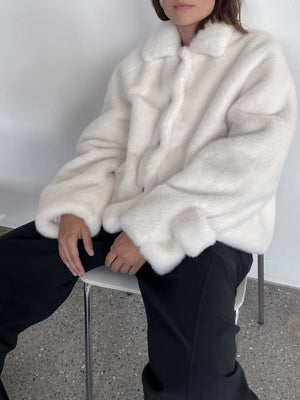 Hvid, fluffy jakke med knapper, bæres af en person, der sidder på en stol. Perfekt til kolde dage fra MEOTINE.