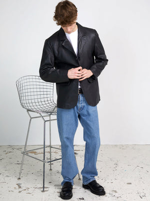 Mand iført en sort læderjakke fra MEOTINE, kombineret med lyse jeans og sorte sko, står ved en moderne barstol.