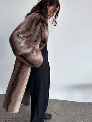 Elegant brun pelsfrakke fra MEOTINE, båret af en kvinde med bølget hår, iført sorte bukser og stående på en grå overflade.