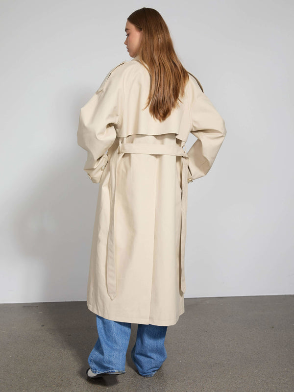 Beige trenchcoat fra MEOTINE set bagfra, med bælte og løse ærmer, kombineret med blå jeans. Elegant og moderne stil.